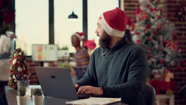 クリスマスの季節を祝う騒々しい人々によって中断される お祝いの装飾されたオフィスで働くことを試みる圧力をかけられた人 お祭り騒ぎの同僚 サンタの帽子を持つ不穏な男 三脚ショット — ストック動画
