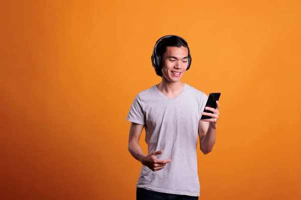 头戴无线耳机的男人在手机上看视频 笑着听播客的亚洲少年 年轻快乐的人 戴着耳机 在智能手机上欣赏音乐 — 图库照片