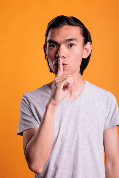 Young asian man showing silence gesture, making tsss sign. Serious teenager loking at camera with forefinger on lips, attractive person keeping secret, studio medium shot