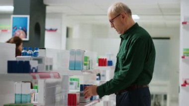 Eczanedeki ilaç kutularına bakan, eczane raflarından ilaç satın alan kıdemli bir müşteri. Yaşlı adam ilaç ve kapsül paketlerini kontrol ediyor, tedavi görüyor..