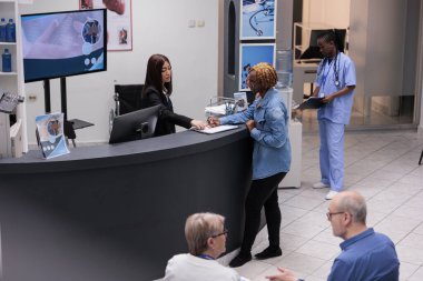 Hastane resepsiyonunda kayıt formunu dolduran genç bir yetişkin randevuya katılmadan önce sağlık kontrolü raporu yazıyor. Lobide evrakları kontrol eden bir kadın var..