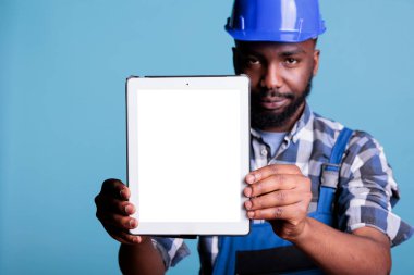 Ciddi inşaat işçisi kameranın önünde boş ekranlı elektronik cihaz tutuyor. Stüdyoda dijital tablet kopyalama alanı olan bir yapıcı mavi arkaplana karşı çekim yapar.