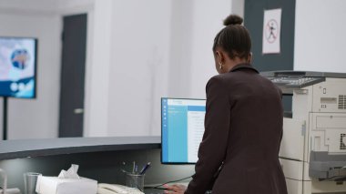 Hastanenin kayıt gişesinde randevuları ve sağlık raporlarıyla çalışan Afrikalı Amerikalı resepsiyonist. Klinik çalışanı danışma masasındaki monitörden sağlık kontrollerine bakıyor..