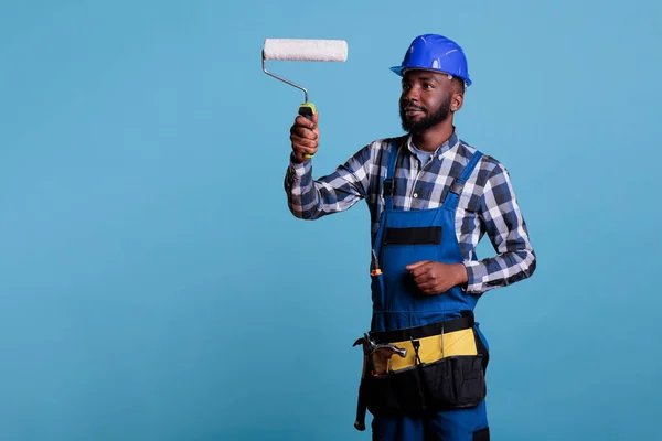 Painter focused on work holding professional roller to paint an interior wall. Construction worker enjoying job dressed in coveralls against blue background, studio portrait.
