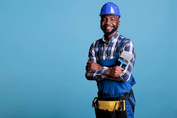 Positive male professional painter standing with brush in hands, wearing work uniform and protective helmet with arms crossed. Interior studio isolated on blue background.
