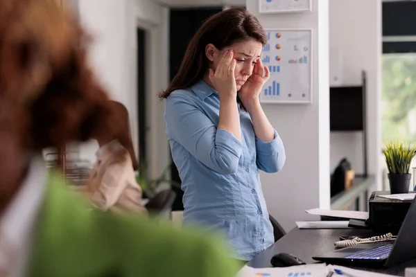 職場の机の近くに立っている同僚のスペースで頭痛 寺院をこすり 疲れ果てた女性を持っている疲れオフィスの従業員 片頭痛の症状に苦しむストレス労働者 — ストック写真