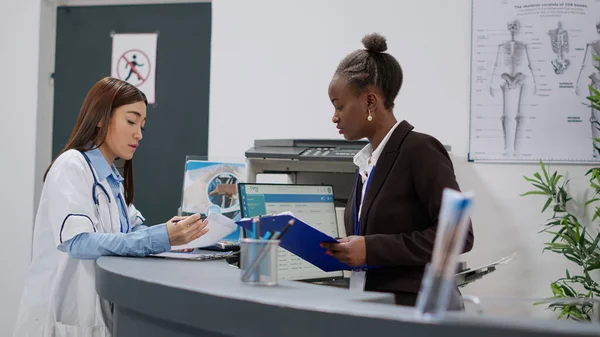 Diverse team of doctor and receptionist working at reception desk to fill in medical report papers and make healthcare appointments. Medical staff doing registration work in facility lobby.