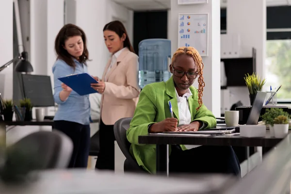 Diverse employees in coworking open space, worker writing on clipboard, sitting at workplace desk. Young african american woman working with company documents, taking notes