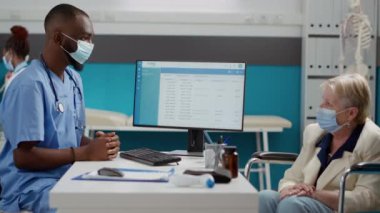 Medical worker talking to woman wheelchair user about disease, dealing with physical impairment. Nurse and senior patient doing checkup consultation in cabinet, wearing face masks.