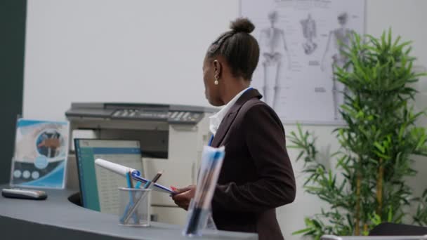 アフリカ系アメリカ人の受付担当者が 健康診断フォームTを使用して紙広告コンピュータ上の医療報告書を確認し 保険サポートで健康保険の予約を行います カウンターで登録作業を行う — ストック動画