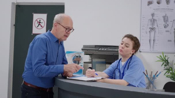 Paciente Sênior Conversando Com Enfermeira Que Trabalha Recepção Hospital Preenchendo — Vídeo de Stock