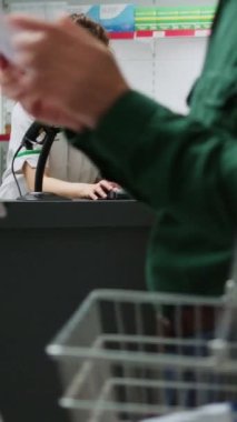 Dikey video: Afrika kökenli Amerikalı polis memuru eczane ürünleri alıyor ve eczane tezgahında banka kartıyla ödeme yapıyor. Temassız ödemelerle ilaç ve sağlık malzemeleri almak için...