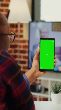 Dikey video: Genç yönetici yeşil ekran ekranlı cep telefonunu dikey olarak tutuyor, merkez ofiste oturuyor. İzole edilmiş krom anahtar şablonu ve boş telif alanı ile çalışmak, arkaplanı düzenle
