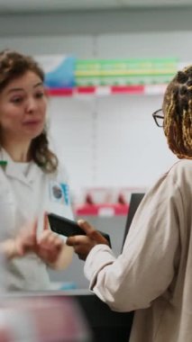 Dikey video: Afrikalı Amerikalı eczacı eczaneden almak için eczacıya ilaç şişesinde reçeteli tedaviyi soruyor. İlaç ürünleriyle kadınlara hizmet eden bir tıp çalışanı.