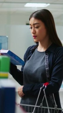 Dikey video: Asyalı müşteri ilaç kutularına ve raflardaki ilaç şişelerine bakıyor, tıbbi ürünler ve takviye malzemeleri almak için eczacıları kontrol ediyor. İlaç paketi broşürü okuyan kadın.