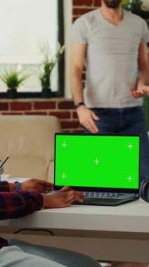 Dikey video: Genç serbest çalışan yeşil ekran ile çalışmak için dizüstü bilgisayar kullanıyor, oturma odasında oturuyor. Boş şablon ile izole edilmiş telif uzayına bakılıyor, kroma tuş görüntüsünü taklit ederek çalışıyor.