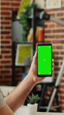Dikey video: Ofis çalışanı yeşil ekran şablonu ile akıllı telefonu analiz ediyor, kroma anahtarı ve kopyalanmış boşluk kullanarak izole görüntü kullanıyor. Kablosuz cep telefonunun boş model geçmişiyle çalışmak.