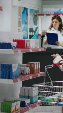 Dikey video: Eczaneden reçeteli ilaç ve vitamin almak için raflardaki ilaçları inceleyen Afrikalı Amerikalı müşteri. Eczane tezgahında oturan müşteri ilaç almak için