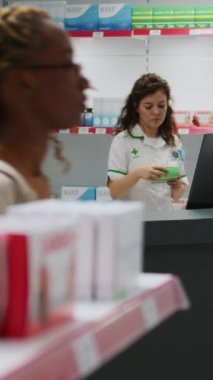 Dikey video: Gülümseyen sağlık çalışanı, hastalık ve reçeteli tedaviye yardımcı olmak için vitamin kutularını işaretliyor. Eczane tezgahında çalışan ve ilaç ve ürünlere bakan eczacı..