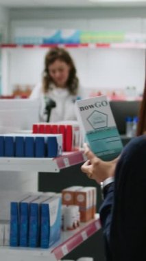 Dikey video: Eczane tezgahındaki Asyalı kadına tıbbi tedavi ve vitaminleri açıklayan sağlık danışmanı ilaç ürünleri paketini gösteriyor. Çeşitli insanlar tıptan bahsediyor..