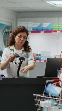 Dikey video: Çeşitli müşteriler eczanede tıbbi ürünleri akıllı sehpayla, kredi kartıyla ve telefonla ödemek için sırada bekliyorlar. Eczacı eczanede ilaç ve takviye malzemeleri satıyor.