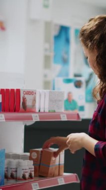 Dikey video: Eczane perakende mağazasında ilaç ve vitamin içeren rafları inceleyen kadın müşteri, ilaç almak için ilave paketlere bakıyor. İlaç ürünleri ve şişeleri kontrol ediliyor