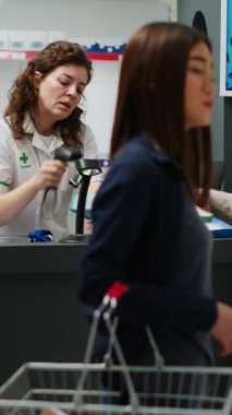 Dikey video: Kadın ilaç ve ilaç ürünlerini ödemek için bankamatik kartını kullanıyor, sağlık sigortası ilaçları almak için temas kurmadan ödeme yapıyor. Eczacı, eczanede insanlara yardım ediyor. Üçayak