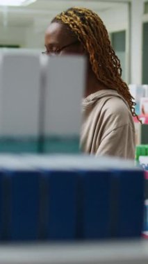 Dikey video: Afro-Amerikan gülümseyen kadın eczane raflarındaki ilaç paketlerini kontrol ediyor, eczanedeki kutularda sağlık ürünleri ve vitaminler alıyor. Hap ve uyuşturucuya bakan kişi.