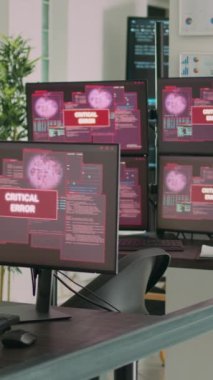 Dikey video: Ekranda hata mesajı gösteren birden fazla monitörlü masa, hacklenen bilgisayar sunucusu. Sistem hatası güvenliği ve hackleme uyarısı olan boş yazılım geliştirme bürosu.