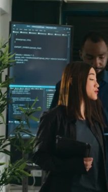 Dikey video: İki yazılım kodlayıcısı dizüstü bilgisayardaki hataları analiz ediyor, ajans ofisindeki büyük televizyon monitörlerini kontrol ediyor. Yoğun şirketlerde siber güvenlik yeniliği üzerinde çalışan programcılar