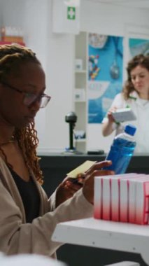 Dikey video: Afro-amerikan müşteriler ilaç ürünleri alıyorlar ve kredi kartıyla ödüyorlar, eczaneden ilaç almak için temas kurmadan ödeme yapıyorlar. Satın almak için eczanede oturuyor.