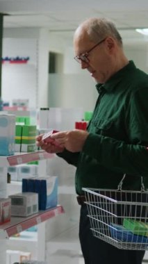 Dikey video: Eczanede gülümseyen ve kardiyoloji hapı kutusuna bakan kıdemli müşteri, reçeteli ilaç ve ilaç almaya hazırlanıyor. İlaçlar ve takviyeler kontrol ediliyor