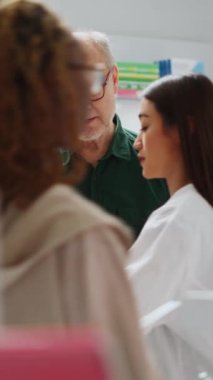 Dikey video: Asyalı eczacı kıdemli müşterilerine ilaç kutularını gösteriyor, hastalık tedavisini ve eczanedeki takviyeleri açıklıyor. Tıp işçisi yaşlı bir adamla ilaç hakkında konuşuyor.