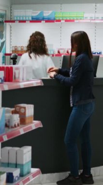 Dikey video: Sağlık uzmanı eczane tezgahındaki Asyalı müşteriye ilaç ve takviyelerden bahsederek hap broşürlerini açıklıyor. Eczacılık ürünleri konusunda müşteriye yardım eden işçiler