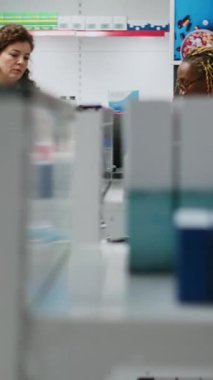 Dikey video: NFC 'li akıllı saat, kredi kartı veya cep telefonu kullanarak eczane masasından ilaç ve takviye malzemeleri alan çok ırklı bir grup insan. İlaç almak için eczane tezgahında ödeme yapmak.