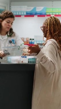 Dikey video: Afrikalı Amerikalı müşteriler eczane tezgahından ilaç ve tedavi satan eczacı ürünleri ve tıbbi paketler alıyorlar. Eczanede ilaç parası ödeyen kişi