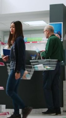 Dikey video: Eczacıyla ilaç tedavisi hakkında konuşan kıdemli müşteri, reçeteli ilaç ve vitamin almak için eczane tezgahında oturuyor. Hastalıklı yaşlı bir adam ilaçlara para ödüyor.