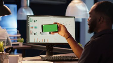 Ofis çalışanı gece geç saatlerde iş ofisinde yatay yeşil ekranlı akıllı telefon tutuyor. İzole görüntüleme ve kopyalama alanı şablonu ile kromakey arkaplanına bakılıyor.