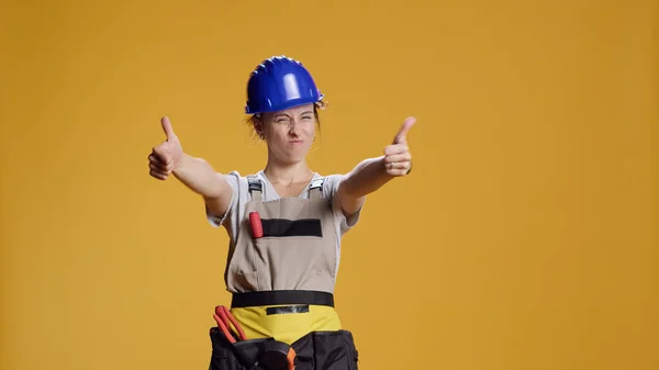 Portrait of female construction worker giving thumbs up in studio, showing approval and agreement on camera. Woman builder doing like and satisfaction gesture, good renovation work.