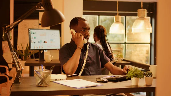 Startup employee using landline phone to chat with people in office during sunset. Answering cord telephone call to talk remotely with manager, working on business report on computer.