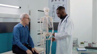 Health specialist analyzing human skeleton bones in cabinet, explaining osteopathy diagnosis to elderly patient. Osteopath examining orthopedic anatomy spinal cord at medical appointment.