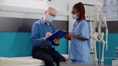 Kıdemli hasta Coronavirus salgını sırasında hemşireyle sağlık kontrolü için form imzalıyor. Yaşlı adam uzman doktorla görüşüyor, reçeteli tedavi görüyor..