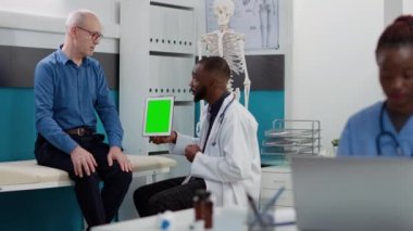 African american medic pointing at tablet with greenscreen display, looking at chromakey template with old patient. Physician and man using isolated mockup background and blank copyspace.