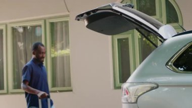 Multiethnic people loading voyage luggage inside car trunk while getting ready for summer holiday citybreak. Diverse tourists going on field trip journey while putting baggage inside vehicle.