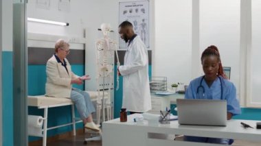 Health specialist examining human skeleton with senior patient in medical cabinet, doing anatomy bones examination, osteopathy spinal cord analysis. Osteopath at checkup visit with old woman.