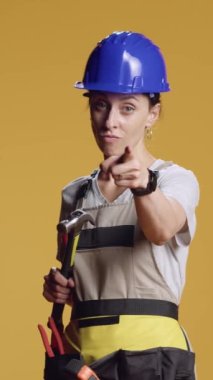 Dikey video: Çekici tutan ve kameraya doğrultan mühendisin portresi, kırıcı aletle agresif olmak. Stüdyoda elinde balyozla işaret etmek için işaret parmağı kullanan kasklı bir kadın..
