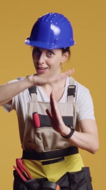 Dikey video: Stüdyoda şekil işareti gösteren kadın müteahhit portresi, inşaat ve dekonstrüksiyon üzerinde çalıştıktan sonra zaman aşımı hareketi yapıyor. İnşaatçı reklam molası ve duraklama, dur sembolü.