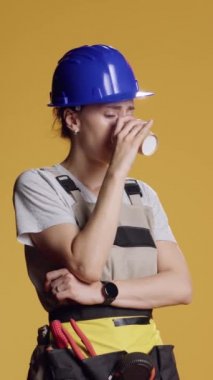 Dikey video: Çalışırken kahve fincanı içen, yorgun hisseden ve uyuyan uykulu kadın portresi. İnşaat işçisi sarı üzerine espresso ve içecek servisi yapıyor.
