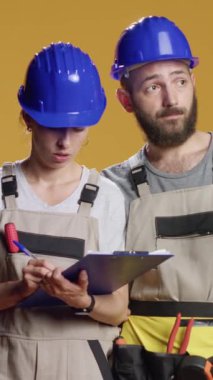 Dikey video: İnşaat işçileri kağıtların ölçümlerini hesaplıyor, notlar alıyor ve inşa sürecini planlıyorlar. Yenileme ve yenilenme ve sanayi sektöründe çalışmak için kask ve tulum giyiyorlar.