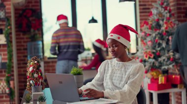 Noel Baba şapkalı Afrikalı kadın laptopla festival ofisine e-posta gönderiyor, kış tatilini iş yerinde kutlamaya hazırlanıyor. Uzayda Noel ağacı ve ışıklarla çalışmak. El kamerasıyla..